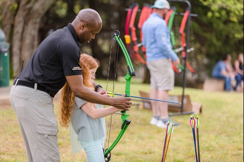 Man helps girl nock an arrow at an archery demonstration